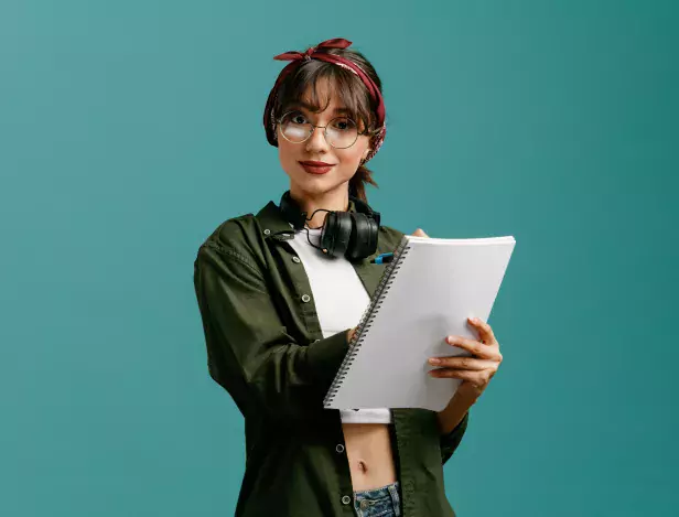 pleased-young-student-girl-wearing-bandana-glasses-and-headphones-around-neck-holding-large-note-pads-looking-at-camera-writing-with-pen-on-note-pad-isolated-on-blue-background